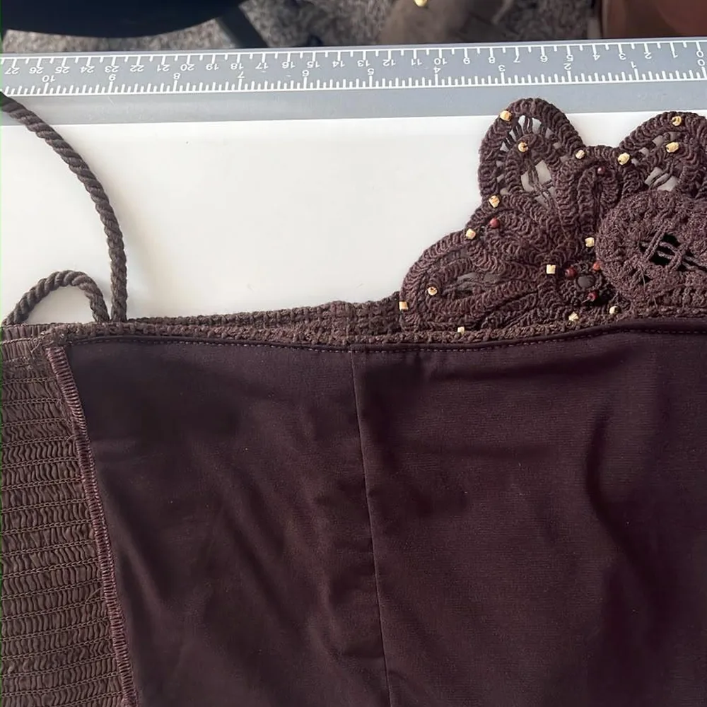 Uma blusa marrom com acabamento de renda de crochê e miçangas douradas, sobre a mesa de corte